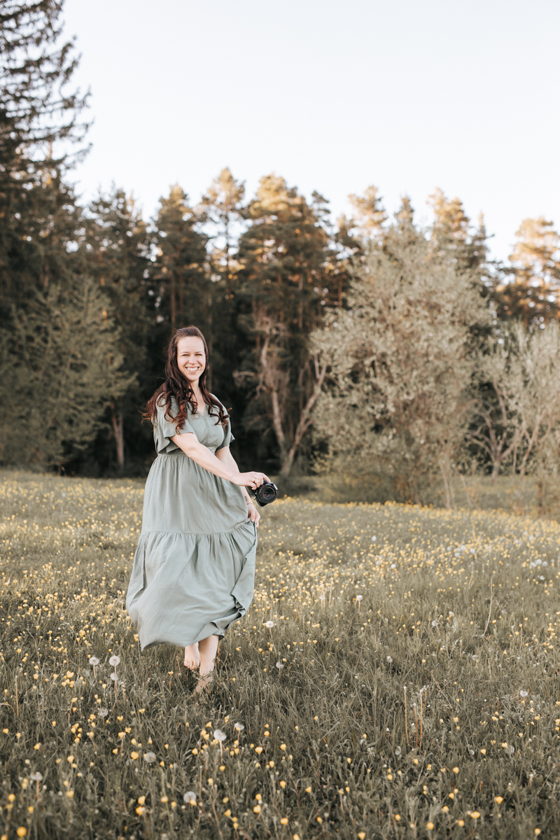 Eine junge Fotografin die Barfuß und im langen Kleid über eine Blumenwiese läuft. Sie lächelt breit.