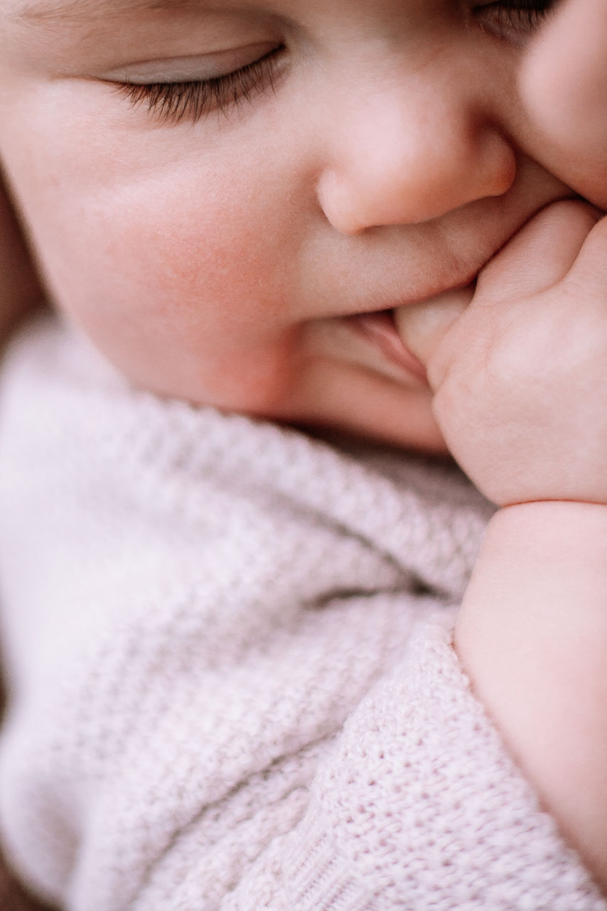 Ein Baby, das an seinem Daumen nuckelt. Es hat die Augen geschlossen und lächelt dabei. 