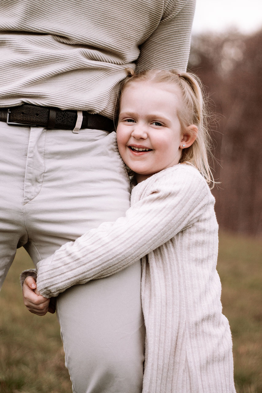 Ein Mädchen das die Hände um Papas Bein geschlungen hat. Sie lacht frech in die Kamera. 