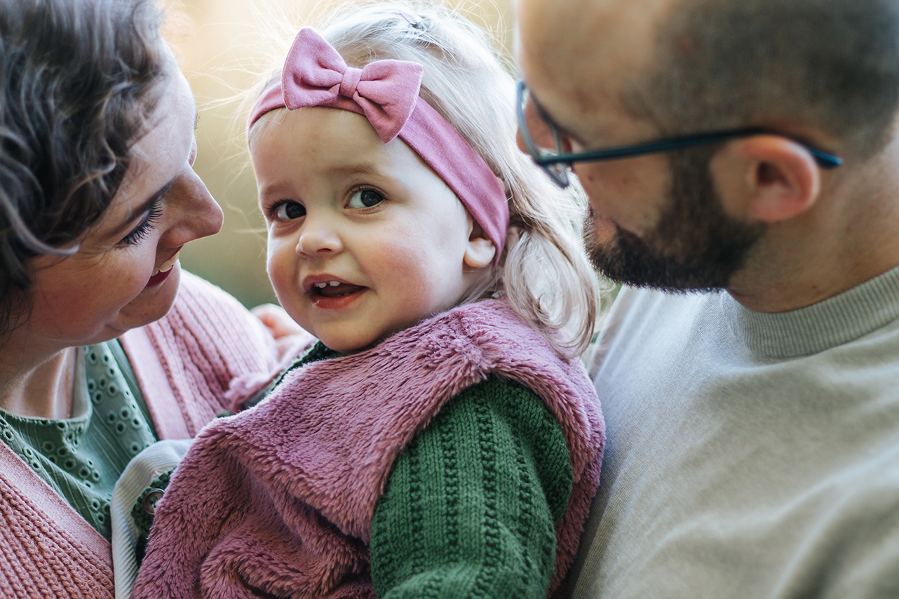 Ein kleines Mädchen in den Armen der Mutter. Der Vater steht hinter dem Mädchen. Beide Eltern lächeln das Kind an. Sie stehen nah beisammen.